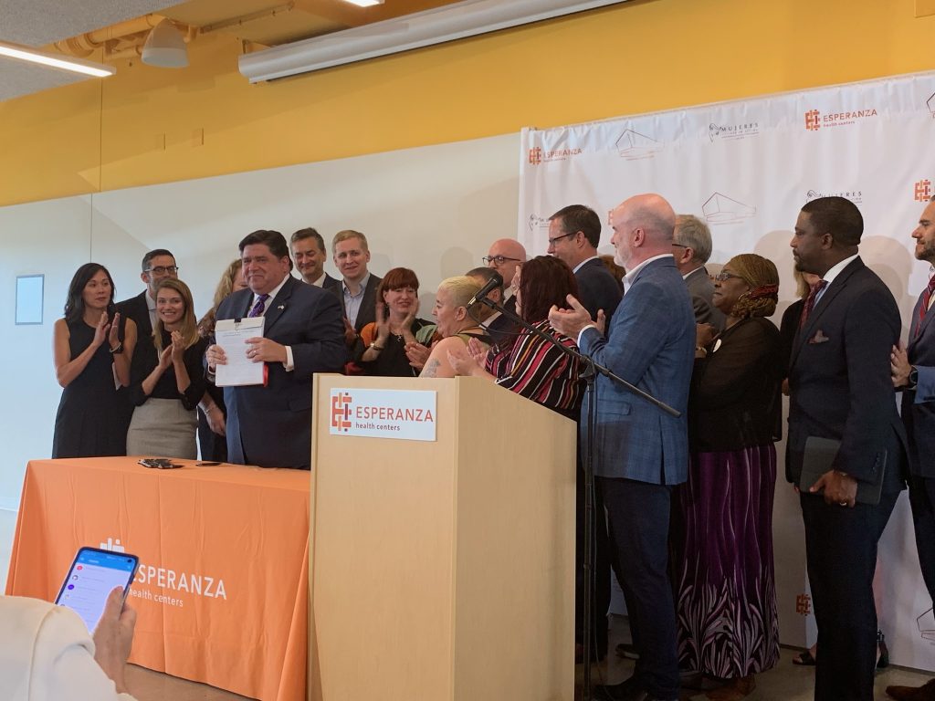 governer pritzker holding a just-signed bill as the people around him applaud.